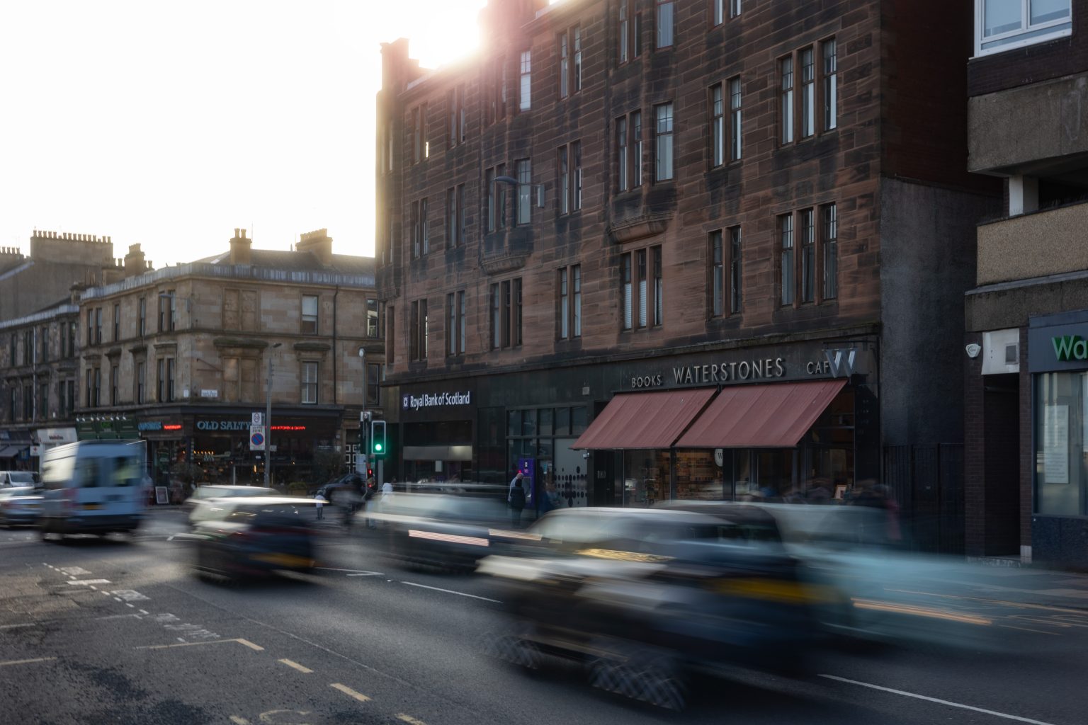 Byres Road, Hillhead, Glasgow, shops, cars whizzing past, blonde and red sandstone tenements