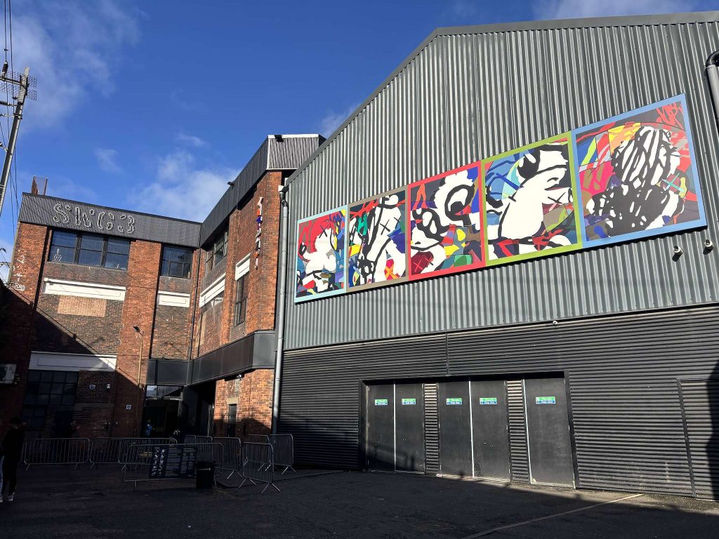 SWG3, Finnieston, Glasgow, a brick warehouse with SWG3 lettering beside a grey clad hall hung with colourful panel artworks